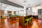 The clubroom at Senate Square is a thoughtfully designed single-floor amenity space that seamlessly blends classic architectural character with modern comforts. Rich hardwood flooring flows throughout, anchoring each distinct zone within the open layout.

The lounge area serves as a focal point, centered around an exposed brick accent wall that frames a sleek fireplace topped with a wall-mounted flat-screen television. Comfortable seating, including leather sofas and upholstered armchairs, is arranged around a glass-top coffee table atop a decorative area rug, creating an inviting gathering space. Multiple large windows along the perimeter flood the room with natural light and offer views of the surrounding greenery.

Adjacent to the lounge, a billiards area features a full-size pool table beneath an array of industrial-style pendant lights. A dark wood bar counter with green-cushioned stools provides a casual spot for socializing, while a flat-screen TV mounted on the brick wall adds to the entertainment options.

The kitchenette is well-appointed with dark cabinetry, granite countertops, stainless steel appliances including a double oven and dishwasher, and a window above the sink. The space is finished with recessed and pendant lighting throughout, contributing to a warm, polished ambiance ideal for community gatherings.