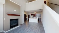 This two-story residence is designed with an open, airy layout that makes a strong first impression. The main living area benefits from dramatically tall ceilings that extend upward toward the second floor, amplifying the sense of space. A sleek fireplace with a wood mantel serves as a focal point, while large sliding glass doors lead to a balcony, flooding the room with natural light and grey-toned hardwood-style flooring throughout.

The kitchen is well-appointed with rich dark cabinetry, light granite-style countertops, stainless steel appliances including a refrigerator, range, and microwave, and a central island that provides additional workspace and casual seating. Striking red pendant lights add a modern accent above the island.

A hallway near the kitchen reveals staircase access to the upper level, where a spacious loft-style area with sloped ceilings and plush carpeting offers versatile use. Bedrooms on both floors feature neutral tones, soft carpet, and ample room. Bathrooms are finished with dark vanity cabinetry, stone-look countertops, and either a walk-in shower with sliding glass doors or a full vanity configuration, reflecting a consistent and polished design throughout.