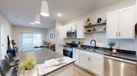 This well-appointed 1-bedroom, 1-bathroom unit offers a thoughtfully designed layout across a single floor, making efficient use of its compact footprint. The open-concept kitchen is a standout feature, showcasing white shaker cabinetry, dark countertops, stainless steel appliances including a gas range, microwave, dishwasher, and refrigerator, as well as open wood shelving for additional storage and display. A large island with seating anchors the space and flows seamlessly into the dining and living areas, all set atop light-toned hardwood-style flooring. The living room is bright and inviting, highlighted by a large sliding glass door that fills the space with natural light and offers outdoor views. The bedroom is a comfortable retreat featuring plush carpet, a natural light-filled window, and access to a dedicated closet. The bathroom is finished with white cabinetry, dark countertops, a tall linen tower, and matte black hardware accents. A dedicated in-unit laundry room with a full-size top-load washer and dryer and built-in wire shelving adds everyday convenience. Neutral tones, recessed lighting, and modern fixtures throughout create a clean, contemporary ambiance.