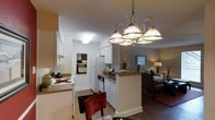 This single-floor residence offers an open-concept layout that seamlessly connects the kitchen, dining, and living areas. The kitchen is outfitted with white cabinetry, a white appliance suite including a range and refrigerator, and a peninsula with dark countertops that provides additional prep space and separates the kitchen from the adjacent areas. A stainless steel dishwasher is also integrated into the kitchen layout.

A decorative multi-arm chandelier with frosted glass shades serves as a focal point above the dining area, adding an ornate touch to the space. The flooring throughout is hardwood-style, contributing to a cohesive and clean aesthetic. Warm, neutral wall tones complement the white trim and cabinetry, creating an inviting atmosphere.

The living area beyond the kitchen is warmly lit, furnished with seating and a side table with a lamp, and features a window that allows natural light to filter in. A textured ceiling is consistent throughout the space. The overall layout promotes an open flow between functional zones, making the space feel connected and practical for everyday living.