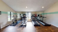 Located at 813 Del Rio Pike in Franklin, TN, this well-equipped fitness center offers a comprehensive workout environment within a single-floor layout. The space is defined by warm hardwood flooring throughout, vaulted and textured ceilings, and a distinctive teal accent stripe that runs along the walls, lending a clean, energetic aesthetic.

The cardio zone is generously stocked with multiple treadmills, elliptical trainers, and stationary bikes, all positioned to take advantage of the large windows that line the walls and flood the space with natural light. A full-length mirror along one wall amplifies the sense of openness and allows users to monitor their form.

The strength training area includes an Inspire cable machine with an attached bench, ideal for a wide range of resistance exercises. A tiered dumbbell rack holds a full set of hex weights, complemented by resistance bands mounted on the wall. A rack of medicine balls and stability balls of varying sizes rounds out the functional training options.

Entry is through double glass-panel doors marked "Fitness Center," creating a welcoming, professional entrance. Additional amenities include sanitation stations with spray bottles and paper towel dispensers, a small storage shelf, and multiple access doors providing convenient flow throughout the space.