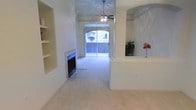 This single-floor residence offers a well-appointed and inviting living area designed with both comfort and functionality in mind. The main living space is anchored by a tiled fireplace set into the wall, accompanied by built-in shelving that provides both display and storage opportunities. Soft, neutral-toned carpet flows throughout, contributing to the warm and airy ambiance of the interior. A ceiling fan is centrally positioned overhead, ensuring year-round comfort while adding a classic architectural touch. The space benefits from generous natural light streaming through vertical-blind-covered sliding glass doors, which lead to an outdoor area and frame views of the exterior environment. A half-wall partition subtly defines distinct zones within the open layout, lending a sense of structure without compromising the openness of the floor plan. The walls are finished in a clean, light tone that enhances brightness throughout. Overall, this space presents a cohesive, functional layout with thoughtful built-in features, ample natural light, and a comfortable atmosphere well-suited for everyday living.