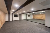 This single-floor gym is designed with a clean, contemporary aesthetic that balances functionality and style. The main workout area is generously proportioned, offering an open floor plan covered in durable speckled rubber flooring suited for a variety of exercise activities. A striking wood-paneled accent wall runs along one side of the space, adding warmth and texture that contrasts with the otherwise sleek, neutral color palette of white walls and dark trim. Full-length mirrors line an entire wall, amplifying the sense of space and allowing users to monitor their form during workouts. A wall-mounted flat-screen display is positioned for clear visibility throughout the room, supporting guided workouts or entertainment. The ceiling features bold dark beam accents with recessed lighting, providing bright, even illumination across the entire floor. Entry to the gym is through glass-paneled doors, adding a polished, professional touch to the space. Additional amenities visible near the entrance include a small bench and storage for personal items. The overall ambiance is modern and motivating, with a well-considered design that supports both individual training and group fitness activities.