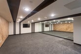 This single-floor gym is designed with a clean, contemporary aesthetic that balances functionality and style. The main workout area is generously proportioned, offering an open floor plan covered in durable speckled rubber flooring suited for a variety of exercise activities. A striking wood-paneled accent wall runs along one side of the space, adding warmth and texture that contrasts with the otherwise sleek, neutral color palette of white walls and dark trim. Full-length mirrors line an entire wall, amplifying the sense of space and allowing users to monitor their form during workouts. A wall-mounted flat-screen display is positioned for clear visibility throughout the room, supporting guided workouts or entertainment. The ceiling features bold dark beam accents with recessed lighting, providing bright, even illumination across the entire floor. Entry to the gym is through glass-paneled doors, adding a polished, professional touch to the space. Additional amenities visible near the entrance include a small bench and storage for personal items. The overall ambiance is modern and motivating, with a well-considered design that supports both individual training and group fitness activities.