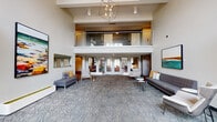 This well-appointed community clubhouse is designed to impress from the moment you enter. The grand lobby greets visitors with soaring double-height ceilings, a striking geometric chandelier, and patterned herringbone carpet that anchors the open, airy layout. A glass-railed mezzanine level overlooks the main floor, adding architectural depth and visual interest throughout the space.

The main lounge area is furnished with modern gray sofas, accent chairs, and side tables, creating comfortable conversation zones beneath vibrant contemporary artwork. Natural light filters in through large glass-panel entry doors, while a glass-enclosed conference or meeting room near the entrance offers a private setting for small gatherings or business needs, equipped with a round table and green upholstered seating.

A secondary lounge area features warm wood flooring, built-in dark cabinetry, a wall-mounted television, and a sleek fireplace flanked by bench seating and framed artwork — a welcoming retreat for relaxed socializing.

The kitchenette is thoughtfully designed with sage green cabinetry, white quartz countertops, a textured tile backsplash, a built-in beverage cooler, and a bar-height counter with seating — well-suited for catered events or casual use. Together, these spaces form a cohesive, modern clubhouse environment built for community connection.