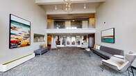 This well-appointed community clubhouse is designed to impress from the moment you enter. The grand lobby greets visitors with soaring double-height ceilings, a striking geometric chandelier, and patterned herringbone carpet that anchors the open, airy layout. A glass-railed mezzanine level overlooks the main floor, adding architectural depth and visual interest throughout the space.

The main lounge area is furnished with modern gray sofas, accent chairs, and side tables, creating comfortable conversation zones beneath vibrant contemporary artwork. Natural light filters in through large glass-panel entry doors, while a glass-enclosed conference or meeting room near the entrance offers a private setting for small gatherings or business needs, equipped with a round table and green upholstered seating.

A secondary lounge area features warm wood flooring, built-in dark cabinetry, a wall-mounted television, and a sleek fireplace flanked by bench seating and framed artwork — a welcoming retreat for relaxed socializing.

The kitchenette is thoughtfully designed with sage green cabinetry, white quartz countertops, a textured tile backsplash, a built-in beverage cooler, and a bar-height counter with seating — well-suited for catered events or casual use. Together, these spaces form a cohesive, modern clubhouse environment built for community connection.