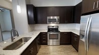 This single-floor space showcases a well-appointed kitchen as a central highlight. The kitchen features rich, dark espresso-toned shaker-style cabinetry that extends from floor to ceiling, providing generous storage throughout. Light-colored granite countertops with subtle veining offer a striking contrast against the dark cabinetry, creating a visually balanced aesthetic. A classic white tile backsplash adds a clean, crisp layer between the upper and lower cabinets.

The kitchen is equipped with stainless steel appliances, including an electric range with a smooth cooktop, a built-in over-the-range microwave, and a stainless steel sink. The appliances are seamlessly integrated into the cabinetry layout, contributing to a cohesive, modern look. The countertops wrap around in a U-shaped configuration, maximizing workspace and functionality.

The flooring consists of light wood-toned planks, lending warmth to the space and complementing the overall color palette. The light-colored walls and neutral tones throughout enhance the brightness of the kitchen. The space presents a clean, move-in-ready environment with modern finishes suited for everyday use.