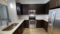 This single-floor space showcases a well-appointed kitchen as a central highlight. The kitchen features rich, dark espresso-toned shaker-style cabinetry that extends from floor to ceiling, providing generous storage throughout. Light-colored granite countertops with subtle veining offer a striking contrast against the dark cabinetry, creating a visually balanced aesthetic. A classic white tile backsplash adds a clean, crisp layer between the upper and lower cabinets.

The kitchen is equipped with stainless steel appliances, including an electric range with a smooth cooktop, a built-in over-the-range microwave, and a stainless steel sink. The appliances are seamlessly integrated into the cabinetry layout, contributing to a cohesive, modern look. The countertops wrap around in a U-shaped configuration, maximizing workspace and functionality.

The flooring consists of light wood-toned planks, lending warmth to the space and complementing the overall color palette. The light-colored walls and neutral tones throughout enhance the brightness of the kitchen. The space presents a clean, move-in-ready environment with modern finishes suited for everyday use.