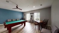 This single-floor recreational space is thoughtfully designed as a social hub for entertainment and leisure. The room is anchored by a classic wood-framed pool table with a green felt surface, positioned centrally to allow ample play space. Light wood-style laminate flooring runs throughout, complementing the neutral gray walls and lending a clean, modern aesthetic to the space.

A bold teal accent wall adds a pop of color to one end of the room, flanked by casual armless lounge chairs — ideal for spectators or those looking to relax. Opposite this, a wall-mounted flat-screen TV sits above a wooden console, paired with a full-size black refrigerator for refreshments. A ceiling fan with integrated lighting keeps the space comfortable.

Natural light pours in through multiple large windows fitted with white horizontal blinds, brightening the room throughout the day. The windowed wall also features bar-height bistro tables and stools, creating an informal gathering spot. Decorative billiards wall art and a chalkboard menu board add personality and charm.

A dedicated game table — featuring a chess and checkers board surface — with surrounding chairs provides an additional activity station. The open layout of this amenity space makes it well-suited for community gatherings, casual socializing, and friendly competition.