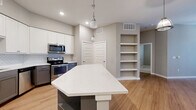 Located at 1300 Camden St in San Antonio, Texas, this single-floor 2-bedroom, 3-bathroom home offers a well-designed layout with modern finishes throughout. The open-concept kitchen and living area serves as the heart of the home, featuring a striking geometric island with a white quartz countertop, white upper cabinetry, two-tone gray lower cabinets, a subway tile backsplash, and stainless steel appliances including a French door refrigerator and dishwasher. Pendant lighting above the island adds a contemporary touch, while warm hardwood-style flooring flows through the main living areas. A built-in open shelving unit provides functional display and storage space adjacent to the kitchen. Both bedrooms are carpeted and receive natural light through windows fitted with blinds; ceiling fans add comfort. The primary bedroom is generously proportioned and includes a well-organized walk-in closet with built-in shelving and hanging rods. The hallways feature additional open shelving, enhancing storage options throughout. A dedicated laundry area houses a stacked Whirlpool washer and dryer unit. The overall palette of soft grays and neutral tones creates a clean, cohesive ambiance across the entire home.