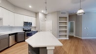 Located at 1300 Camden St in San Antonio, Texas, this single-floor 2-bedroom, 3-bathroom home offers a well-designed layout with modern finishes throughout. The open-concept kitchen and living area serves as the heart of the home, featuring a striking geometric island with a white quartz countertop, white upper cabinetry, two-tone gray lower cabinets, a subway tile backsplash, and stainless steel appliances including a French door refrigerator and dishwasher. Pendant lighting above the island adds a contemporary touch, while warm hardwood-style flooring flows through the main living areas. A built-in open shelving unit provides functional display and storage space adjacent to the kitchen. Both bedrooms are carpeted and receive natural light through windows fitted with blinds; ceiling fans add comfort. The primary bedroom is generously proportioned and includes a well-organized walk-in closet with built-in shelving and hanging rods. The hallways feature additional open shelving, enhancing storage options throughout. A dedicated laundry area houses a stacked Whirlpool washer and dryer unit. The overall palette of soft grays and neutral tones creates a clean, cohesive ambiance across the entire home.