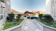 Located at 254 Crown St in the heart of New Haven, CT, this thoughtfully designed outdoor courtyard offers a generous and well-appointed communal space. The grounds are organized into distinct zones that balance greenery and hardscape seamlessly. A broad tiled pathway runs through the center of the space, flanked by raised concrete planters filled with manicured shrubs, flowering plants, and ornamental Japanese maple trees that provide striking seasonal color. The open turf lawn area is lined with vibrant blue Adirondack chairs, creating an inviting spot for relaxation beneath open skies. A dedicated lounge and dining zone features wicker sectional sofas with teal cushions alongside modern green dining chairs and a gas grill, ideal for outdoor gatherings. Horizontal wood-slatted privacy fencing defines the perimeter, offering a sense of enclosure while maintaining an airy atmosphere. The surrounding multi-story building frames the courtyard with large windows and private balconies that overlook the grounds. Views of the broader New Haven skyline are visible beyond the fencing, grounding this outdoor retreat within its urban context. The overall design strikes a balance between contemporary aesthetics and comfortable, functional outdoor living.