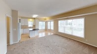 This single-floor residence offers a thoughtfully designed layout that balances functionality and comfort throughout its interconnected spaces. The kitchen serves as the heart of the home, showcasing white cabinetry with glass-front upper panels, warm-toned countertops, and a full suite of white appliances including a range with overhead microwave and dishwasher. A standout central island topped with dark granite adds a striking contrast and provides additional prep and seating space. Natural light pours in through multiple windows in the adjacent dining area, which is accented by pendant lighting overhead. The open living area flows seamlessly from the kitchen, featuring light hardwood flooring that transitions into plush carpeting throughout the bedrooms and hallways. The bedrooms are generously proportioned, with neutral walls, carpet flooring, and ample closet space including built-in shelving. The full bathroom is finished with tile flooring, a soaking tub with tile surround, and a clean white vanity. A dedicated laundry room houses a full-size side-by-side washer and dryer beneath white overhead cabinetry, adding notable convenience. Throughout the space, warm beige and cream tones create a cohesive, welcoming ambiance.