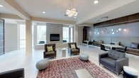 This single-floor amenity space offers a sophisticated, contemporary design with multiple distinct zones for relaxation and entertainment. The entrance features frosted double glass doors with polished chrome hardware, flanked by framed artwork along neutral-toned walls and light tile flooring that flows throughout.

The main lounge area is expansive and open, with recessed lighting and a striking cluster chandelier overhead. Comfortable dark upholstered armchairs are arranged around a glass coffee table atop a patterned area rug, while built-in banquette seating lines one wall, accented by decorative wall-mounted mirrors and soft sconce lighting. A ping-pong table serves as a central recreational feature within the lounge's game zone.

The bar area is a standout highlight, featuring a countertop with integrated LED lighting that casts a vibrant purple glow. Dark granite counters sit atop sleek cabinetry, complemented by a mosaic tile backsplash, a built-in beverage refrigerator, a sink, and multiple wall-mounted flat-screen televisions. Pendant lighting above the bar area adds warmth, while modern bar stools complete the social setting. Overall, the space exudes a polished, upscale ambiance suited for community gatherings.