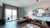 This well-appointed studio offers a thoughtfully designed open-concept layout that seamlessly blends living and sleeping areas into a single, cohesive space. Hardwood-style flooring runs throughout, providing a warm and contemporary foundation that complements the soft, neutral wall tones.

The sleeping area is anchored by a platform-style bed dressed in layered bedding and accent pillows, positioned to take full advantage of the floor-to-ceiling sliding glass doors that lead to a private balcony. The balcony offers sweeping views of the surrounding cityscape, flooding the interior with generous natural light throughout the day. Linen-style curtains frame the doors, adding a soft, elegant touch.

A ceiling fan overhead provides both function and style, while a warm-toned floor lamp and curated gallery wall featuring framed black-and-white photography add character and depth to the space. The living area is defined by a rich, tufted leather sofa paired with a round marble-top coffee table, creating an inviting lounge setting.

Decorative accents — including a small sculptural bicycle figurine, a vibrant floral arrangement, and layered area rugs — contribute to the space's modern yet lived-in charm. Overall, this studio presents a refined urban retreat with a cohesive, design-forward aesthetic.