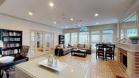 This well-appointed single-floor space offers an open, airy layout that seamlessly connects the living, dining, and kitchen areas into one cohesive environment. The living area is a standout, featuring vaulted ceilings with a striking arched transom window that floods the room with natural light. A ceiling fan, recessed lighting, and a wall of large windows with vertical blinds further enhance the bright, welcoming atmosphere. Comfortable neutral-toned sofas are arranged around a dark wood coffee table, creating an inviting seating area complemented by a freestanding bookcase.

The dining area is positioned adjacent to the living space and is furnished with a rich, dark wood table and matching chairs, with an outdoor view provided through floor-to-ceiling windows. A brick-accented fireplace detail adds warmth and character to this zone.

The kitchenette is efficiently designed with dark upper cabinetry, a countertop with a sink, a microwave, and a prominent white island that serves as both a workspace and a visual anchor for the space. A decorative bench nearby adds a thoughtful finishing touch.

Throughout the space, warm-toned herringbone-patterned tile flooring ties the rooms together, while elegant French doors with glass panes provide access to an adjoining area, adding both functionality and architectural charm.