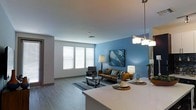 This well-appointed single-floor residence offers a thoughtfully designed layout that balances modern aesthetics with everyday functionality. The entryway leads into a long, well-lit hallway finished with light wood-style flooring and striking blue-and-white striped accent walls. Recessed ceiling lights illuminate the corridor, while a sleek sliding barn door adds a contemporary architectural detail and serves as a practical space-saving solution.

The hallway opens into a spacious, open-concept living and dining area that feels bright and airy. Large windows dressed with white blinds allow generous natural light to fill the room, enhancing the welcoming ambiance. A bold blue feature wall serves as a focal point in the living area, providing a vibrant contrast to the neutral tones of the surrounding walls and light flooring. The living space is generously proportioned, offering ample room for comfortable seating arrangements.

The kitchen flows seamlessly into the living area, anchored by a wide, light-toned countertop island that doubles as a casual dining or entertaining surface. Pendant lighting above the island adds a refined touch. Together, the spaces create a cohesive, modern environment that is both functional and visually inviting, ideal for everyday living and hosting.