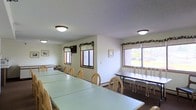This single-floor space is designed as a communal gathering or dining area, offering a practical and welcoming environment suited for group meals, meetings, or events. The room is furnished with several long rectangular tables paired with light wood chairs, arranged to accommodate a significant number of occupants comfortably. The layout is open and organized, allowing for easy movement throughout the space.

Natural light filters in through multiple windows fitted with horizontal blinds and accented with decorative greenery garlands, lending a subtle warmth to the otherwise neutral interior. The walls are painted in a clean, off-white tone, contributing to the bright and airy atmosphere. Additional illumination is provided by flush-mount ceiling light fixtures set against a textured ceiling.

Along one wall, a row of bench-style seating with accompanying narrow tables provides an alternative seating arrangement, adding versatility to the space. Framed artwork is placed on the walls, offering simple decorative touches. The dark flooring contrasts with the light furnishings, grounding the room visually. Overall, the space presents a clean, functional character well-suited for community gatherings.