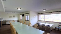 This single-floor space is designed as a communal gathering or dining area, offering a practical and welcoming environment suited for group meals, meetings, or events. The room is furnished with several long rectangular tables paired with light wood chairs, arranged to accommodate a significant number of occupants comfortably. The layout is open and organized, allowing for easy movement throughout the space.

Natural light filters in through multiple windows fitted with horizontal blinds and accented with decorative greenery garlands, lending a subtle warmth to the otherwise neutral interior. The walls are painted in a clean, off-white tone, contributing to the bright and airy atmosphere. Additional illumination is provided by flush-mount ceiling light fixtures set against a textured ceiling.

Along one wall, a row of bench-style seating with accompanying narrow tables provides an alternative seating arrangement, adding versatility to the space. Framed artwork is placed on the walls, offering simple decorative touches. The dark flooring contrasts with the light furnishings, grounding the room visually. Overall, the space presents a clean, functional character well-suited for community gatherings.