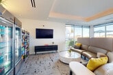 Located at 260 E Rio Salado Pkwy in Tempe, Arizona, this contemporary lounge and dining space offers a thoughtfully designed environment for relaxation and casual dining. The single-floor layout is defined by clean white walls, a tray ceiling with warm ambient cove lighting, and recessed fixtures that create a polished, modern atmosphere throughout.

The lounge area centers around a curved, high-back gray sectional sofa accented with vibrant yellow throw pillows, arranged around a round marble coffee table. Complementary accent chairs provide additional seating, all positioned atop a textured, abstract-patterned area rug. A wall-mounted flat-screen television anchors one side of the room, flanked by a sleek dark media console.

Adjacent to the lounge, a dining area features high-top round tables with woven rattan and aluminum bar-height chairs, ideal for casual dining or socializing. Floor-to-ceiling sliding glass windows and doors line the perimeter, bathing the space in abundant natural light and offering open views of the surrounding outdoor terrace and cityscape.

A built-in snack and beverage station, branded "SALT," provides glass-door refrigerators stocked with drinks and fully loaded snack shelving, adding a convenient amenity within the space. The overall design balances comfort and functionality with a bright, upscale aesthetic suited for community gathering.