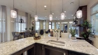 This well-appointed single-floor space is designed with an open-concept layout that seamlessly connects multiple functional zones. The kitchen area anchors the space with a granite-topped island featuring dark cabinetry, a stainless steel sink, and elegant pendant lighting suspended from the ceiling. Recessed lighting throughout ensures the space remains bright and welcoming.

The open layout flows naturally into a comfortable lounge area furnished with neutral-toned sofas and accent chairs, creating an inviting setting for relaxation or casual gatherings. A dining area with upholstered chairs sits nearby, benefiting from abundant natural light streaming through large windows dressed with sheer curtains. Mirrored wall panels enhance the sense of depth and openness throughout the space.

A dedicated workstation area provides a practical counterpoint to the social zones, featuring a granite-topped desk equipped with two desktop computers positioned in front of a large window with slatted blinds, offering outdoor views and natural daylight. Decorative plants are thoughtfully placed throughout, adding warmth and a touch of greenery to the contemporary, neutral color palette.