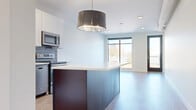 This thoughtfully designed single-floor residence showcases a contemporary open-concept layout that seamlessly connects the kitchen and living areas. The kitchen is a standout feature, centered around a substantial island with a crisp white countertop and rich dark cabinetry, complemented by a bold drum pendant light overhead. White upper cabinets, a slate-tiled backsplash, and a full suite of stainless steel appliances complete the space. Light wood-look flooring flows throughout, enhancing the bright, airy atmosphere created by large floor-to-ceiling windows that flood the living area with natural light.

A private balcony with modern metal railings offers open city views, adding an urban appeal to the residence. The bedroom is generously proportioned, featuring the same expansive windows and light flooring, alongside a walk-in closet outfitted with wire shelving systems and carpeted flooring for comfort. A secondary carpeted room provides flexible use as a bedroom or office.

The two bathrooms are finished in a clean, modern style. One features a soaking tub with full-height textured tile surround and light floor tiles, while the other offers a dark-stained vanity with white countertop, undermount sink, and a large mirror.
