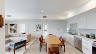 This modern single-floor unit offers five bedrooms and one bathroom within a thoughtfully designed, contemporary layout. The open-concept common area seamlessly integrates the living room, dining area, and kitchen into one cohesive space. The kitchen is outfitted with stainless steel appliances — including a gas range, built-in microwave, dishwasher, and French door refrigerator — complemented by sleek white cabinetry and clean countertops. A large oval wood dining table anchors the dining zone, providing ample seating for residents. The living room features a wall-mounted flat-screen TV, a sliding glass door that opens to a private balcony, and an abundance of natural light. The five bedrooms maintain the unit's minimalist aesthetic, with crisp white walls, large windows with curtains, and compact yet functional layouts. The primary bedroom offers a notably generous footprint compared to the other rooms. The shared bathroom is finished with stone-toned tile, a floating vanity with brushed gold fixtures, open shelving, and a large mirror. Throughout the unit, light-toned flooring, recessed lighting, and a neutral color palette create a clean, cohesive, and contemporary ambiance.