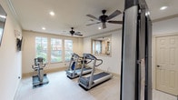 This single-floor fitness center offers a clean, well-organized workout environment designed to accommodate a range of exercise needs. The space is outfitted with multiple treadmills and stationary bikes positioned near large windows that allow natural light to fill the room and provide views of the surrounding outdoor area, creating an inviting atmosphere for cardio workouts. Ceiling fans are distributed throughout the space, working in tandem with recessed lighting to maintain a comfortable and well-lit environment.

The strength training area features Body-Solid multi-function machines, positioned on protective floor mats to define the workout zones. Wall-mounted flat-screen televisions are installed at multiple points throughout the room, offering entertainment during exercise sessions. The light-toned flooring features a subtle tile pattern that contributes to the bright and airy feel of the space.

A dedicated alcove near the entrance houses a wall-mounted water and sanitation station with a built-in shelf, offering added convenience for users. The neutral color palette of cream and beige tones throughout the walls and trim provides a calm, focused atmosphere. The overall layout is thoughtfully arranged to allow users to move freely between cardio and strength equipment without obstruction.