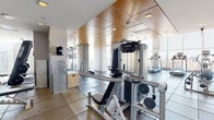 This high-rise fitness center offers a comprehensive and modern workout facility designed to meet a wide range of exercise needs. The space is organized into distinct functional zones, beginning with a cardio area lined with Precor treadmills and elliptical machines positioned directly in front of expansive floor-to-ceiling windows. These large windows flood the space with natural light and frame impressive panoramic views of the surrounding cityscape and waterway below, making for an inspiring workout environment. The flooring throughout transitions between large-format tile and rubberized gym matting, providing both durability and comfort underfoot. A dedicated strength training area houses a variety of Precor cable and resistance machines, along with a dumbbell rack and functional training equipment including a plyo box. The ceilings in the strength zone are finished with warm wood paneling accented by recessed lighting, lending a refined aesthetic contrast to the industrial nature of the equipment. A standout architectural feature is a sleek spiral staircase, finished in brushed metal, which serves as a sculptural focal point within the space. Built-in storage lockers are conveniently accessible near the staircase. The overall design balances functionality with upscale finishes, creating a fitness environment that is both practical and visually distinctive.
