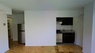 This single-floor space offers a clean, open layout that maximizes functionality and ease of movement. The kitchen is a standout feature, outfitted with dark espresso cabinetry that provides a bold contrast against light granite countertops. A stainless steel gas range and refrigerator complete the cooking area, lending a modern, utilitarian aesthetic. A partial wall separates the kitchen from the adjacent living area, creating a subtle distinction between the two zones while maintaining an open, airy feel. The living area features warm parquet hardwood flooring in a classic block pattern, adding texture and character to the space. The walls are finished in a neutral white throughout, creating a bright and versatile backdrop that complements a variety of décor styles. The overall ambiance is practical and understated, with a layout designed for efficient everyday living. The seamless transition between the kitchen and living area makes the space feel cohesive and well-proportioned, ideal for those who value a straightforward, functional home environment.