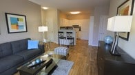 This single-floor residence offers a thoughtfully designed open-concept layout that seamlessly connects the living area to the kitchen, creating a spacious and inviting atmosphere. The living room is anchored by a sleek glass-top coffee table and a comfortable sofa accented with decorative throw pillows, complemented by a stylish table lamp that adds warm ambient lighting. A striped accent chair and matching ottoman provide additional seating and a touch of classic charm.

The hardwood-style flooring runs throughout the main living areas, lending warmth and continuity to the space. Neutral-toned walls enhance the bright, airy feel, further supported by flush-mount ceiling lighting.

The kitchen is efficiently laid out with ample white cabinetry extending to the upper walls, offering generous storage. A dark tile backsplash provides a striking contrast to the light countertops and cabinetry. A peninsula-style breakfast bar with two bar stools serves as both a functional dining spot and a natural divider between the kitchen and living areas.

Multiple doorways visible from the main living space suggest access to additional private rooms, offering a well-organized and functional floor plan throughout this comfortable single-level home.