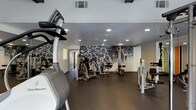 This single-floor fitness center offers a comprehensive workout environment designed to accommodate a range of exercise needs. The space is outfitted with a diverse selection of equipment, including cardio machines such as ellipticals and steppers, as well as multiple strength training stations and cable machines, providing users with full-body workout options. The flooring is a durable dark rubber material, standard for high-traffic fitness environments, offering both safety and functionality underfoot. Recessed lighting is evenly distributed across the ceiling, ensuring the space is well-lit throughout. A standout design element is the expansive black-and-white photographic mural covering an entire wall, depicting a dynamic athletic or cycling event crowd scene, lending the space an energetic and motivating ambiance. The neutral white walls complement the monochromatic mural, keeping the aesthetic clean and focused. A doorway visible at the rear of the space suggests access to additional rooms or facilities. Overall, the fitness center strikes a balance between practicality and visual interest, creating an inviting and functional environment suited for regular, dedicated use.
