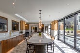 Located at 2333 Calle Del Mundo in Santa Clara, CA, this thoughtfully designed single-floor amenity space offers a refined blend of function and style across a generous footprint. The living and lounge area welcomes visitors with plush, curved sofas, cane-accented armchairs, and a dark marble coffee table, all set beneath a tray ceiling with recessed lighting and warm sconce fixtures. Arched full-length mirrors and woven wall panels add artistic depth to the bright, open atmosphere, while large glass doors invite natural light and outdoor views.

The dining area showcases dramatic curved booths lined with textured gold tile walls and ambient underlighting, paired with marble-top tables and pendant lighting — creating an intimate yet sophisticated setting. A second dining zone features a long, sculptural oval island with a white stone countertop, set atop a striking geometric-patterned tile floor. Rich walnut cabinetry, integrated appliances, pendant lights, and a wall-mounted display screen complete this gourmet communal kitchen.

A dedicated media lounge offers built-in booth seating with warm wood slat paneling, a large wall-mounted screen, and marble wall accents — ideal for both collaborative gatherings and relaxed viewing. Throughout, the space maintains a cohesive, upscale aesthetic with neutral tones, natural materials, and curated furnishings.