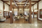 This single-story space makes a striking first impression with a grand entry foyer showcasing ornate wrought-iron double doors beneath a sweeping arched transom, flanked by warm sconce lighting and travertine tile flooring set in a classic diamond pattern. The soaring ceilings throughout create an open, airy ambiance, accentuated by bold dark wood columns, coffered trim details, and an elegant wrought-iron chandelier that serves as a central focal point.

The main gathering area is anchored by a dramatic floor-to-ceiling stacked stone fireplace with a substantial wood mantel, lending the space a rich, lodge-inspired character. Deep leather sofas and an ornately carved coffee table are arranged on a plush area rug, creating a comfortable and inviting lounge setting. Hardwood flooring flows throughout, complementing the dark wood millwork that frames each architectural element.

Adjacent to the lounge, a private conference or meeting room is accessible through full-lite French doors with dark wood framing, offering a functional yet refined space. A kitchenette area with dark wood cabinetry is also visible, adding everyday convenience. Wall sconces provide warm, layered lighting throughout, reinforcing the cohesive, sophisticated aesthetic of this elegantly designed space.