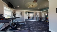 This single-floor fitness center is thoughtfully designed to provide a comprehensive workout environment in a clean, modern setting. The space is anchored by dark, patterned carpet flooring that adds a sleek aesthetic throughout. The walls are finished in a neutral gray tone, accented by a teal-painted ceiling that lends the room a distinctive, contemporary character.

The cardio zone is well-appointed with multiple Matrix-brand treadmills, an elliptical trainer, and a stair climber, all positioned near large windows that invite generous natural light and offer views of the surrounding outdoor greenery. Wall-mounted flat-screen TVs are positioned above the cardio equipment for entertainment during workouts.

On the strength training side, the space includes a multi-function cable and weight machine, a dumbbell rack, and additional resistance equipment. Full-length wall mirrors visually expand the space and allow users to monitor their form during exercise. Decorative abstract wall art adds a polished, upscale touch throughout.

Recessed lighting and a ceiling fan with a brass fixture provide both practical illumination and comfortable airflow. A water cooler station and fire safety equipment are conveniently located along one wall. Multiple windows on opposing walls ensure the space remains bright, airy, and connected to the outdoors throughout the day.