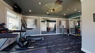 This single-floor fitness center is thoughtfully designed to provide a comprehensive workout environment in a clean, modern setting. The space is anchored by dark, patterned carpet flooring that adds a sleek aesthetic throughout. The walls are finished in a neutral gray tone, accented by a teal-painted ceiling that lends the room a distinctive, contemporary character.

The cardio zone is well-appointed with multiple Matrix-brand treadmills, an elliptical trainer, and a stair climber, all positioned near large windows that invite generous natural light and offer views of the surrounding outdoor greenery. Wall-mounted flat-screen TVs are positioned above the cardio equipment for entertainment during workouts.

On the strength training side, the space includes a multi-function cable and weight machine, a dumbbell rack, and additional resistance equipment. Full-length wall mirrors visually expand the space and allow users to monitor their form during exercise. Decorative abstract wall art adds a polished, upscale touch throughout.

Recessed lighting and a ceiling fan with a brass fixture provide both practical illumination and comfortable airflow. A water cooler station and fire safety equipment are conveniently located along one wall. Multiple windows on opposing walls ensure the space remains bright, airy, and connected to the outdoors throughout the day.
