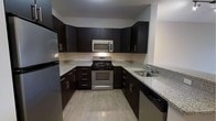 This single-floor space offers a thoughtfully designed layout that balances comfort and functionality. The kitchen is a standout feature, finished with rich dark cabinetry, granite-look countertops, and a full suite of stainless steel appliances including a gas range, over-the-range microwave, refrigerator, and dishwasher. The U-shaped kitchen configuration maximizes workspace and storage, making it well-suited for everyday cooking. Light wood-tone flooring runs through the kitchen, contributing to a clean, contemporary aesthetic.

The floor plan, as revealed in the Matterport dollhouse view, includes a generous open living area that benefits from multiple windows along the exterior walls, allowing natural light to flow freely through the space. The layout also features two bathrooms, each equipped with essential fixtures, providing convenience for residents and guests alike. Interior hallways connect the various zones of the unit efficiently, ensuring smooth transitions between living, sleeping, and utility areas.

Overall, the space presents a modern, neutral palette with consistent finishes throughout, creating a cohesive and inviting atmosphere. The combination of practical storage solutions, quality surface materials, and well-distributed natural light makes this a highly functional and comfortable single-floor living space.