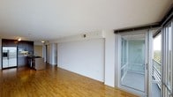This single-floor space offers a clean, modern layout designed for comfortable everyday living. The main living area is characterized by smooth white walls, warm-toned hardwood flooring, and a generous sense of openness that makes the space feel airy and inviting. Natural light filters in through a full-height sliding glass door on one side, which opens directly onto an enclosed balcony — an ideal spot for relaxing or enjoying fresh air. The transition between indoor and outdoor living is seamless, with the balcony offering additional usable space beyond the main interior. Toward the back of the living area, the space flows naturally into a kitchen, partially visible from the main room. The kitchen features dark countertops and cabinetry, providing a subtle contrast to the light tones found throughout the rest of the space. Overhead, the smooth white ceilings contribute to the bright and open atmosphere. Ventilation is provided via ceiling-mounted HVAC vents, ensuring year-round comfort. The overall aesthetic is minimalist and contemporary, with neutral finishes and a practical layout that offers flexibility for a variety of furnishing arrangements. The space is well-suited for those seeking a clean, functional, and light-filled environment.