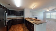 This single-floor residence offers a well-designed open layout that connects the kitchen and living areas seamlessly. The kitchen is a standout space, featuring rich dark-espresso cabinetry with brushed nickel hardware, stone-look countertops, a double-basin stainless steel sink set into a peninsula, and hardwood flooring. The peninsula provides additional workspace while creating a natural divide between the kitchen and the adjacent living area.

The living room is generously proportioned and highlighted by a tray ceiling with crown molding and a ceiling fan. A tile-surround fireplace anchors one wall, while large windows and a sliding glass door with vertical blinds invite abundant natural light and outdoor views. Plush neutral carpet flows throughout the living and sleeping areas, contributing to a warm, comfortable atmosphere.

The bedroom is a calm, carpeted retreat with a natural-light window and direct access to a closet and the full bathroom. The bathroom features hardwood flooring, a stone-topped vanity with a large mirror, a soaking tub with white tile surround, and neutral gray walls throughout. The textured ceilings are consistent across the home, adding a classic residential character to every room.
