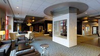 This elegant single-floor space exudes a polished, upscale atmosphere designed for comfort and visual sophistication. At the heart of the room stands a striking floor-to-ceiling column clad in white marble, housing a see-through glass fireplace that serves as a dramatic focal point and natural divider of the space. The patterned carpet underfoot adds texture and visual interest, complementing the rich tones of the leather and upholstered seating arrangements thoughtfully positioned throughout the area. Overhead, a circular recessed ceiling detail crowns the marble column, adding architectural depth alongside a suspended drop ceiling with recessed lighting. Warm, ambient illumination is provided by ornate gold chandeliers that lend a classic, glamorous touch to the contemporary design. The darker accent wall in the background features built-in shelving with backlit displays, creating a moody, lounge-like corner. Sheer curtains and accent lamps further soften the space, while silver side tables add a modern metallic contrast. The overall design balances classic elegance with contemporary flair, creating an inviting and visually dynamic environment suited to hospitality or upscale social gatherings.