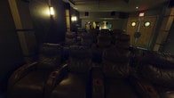 This dedicated movie theater is thoughtfully designed to deliver an immersive cinematic experience. The screening room features multiple tiered rows of oversized, dark leather recliner seats, each equipped with built-in cup holders for added comfort during viewings. The seating arrangement ensures unobstructed sightlines from every position in the room.

The interior is finished in deep, dark tones — including dark walls and rich carpeting with an ornate pattern near the front — creating a true theater ambiance that minimizes light distraction. Decorative column accents with lighter paneling are positioned along the sides of the room, adding architectural interest and housing acoustic panels that contribute to the room's sound quality.

Ambient lighting is provided by elegant wall sconces mounted throughout the space, casting a warm, subdued glow that sets the mood without compromising the viewing environment. A ceiling-mounted projector directs onto a large, dedicated projection screen at the front of the room. Multiple surround sound speakers are strategically placed along the walls and ceiling to ensure a full, enveloping audio experience.

The rear of the theater features double entry doors with porthole-style windows, a classic nod to traditional cinema design, alongside clearly marked exit signage. Overall, the space is purpose-built for premium, private film viewing.