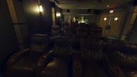 This dedicated movie theater is thoughtfully designed to deliver an immersive cinematic experience. The screening room features multiple tiered rows of oversized, dark leather recliner seats, each equipped with built-in cup holders for added comfort during viewings. The seating arrangement ensures unobstructed sightlines from every position in the room.

The interior is finished in deep, dark tones — including dark walls and rich carpeting with an ornate pattern near the front — creating a true theater ambiance that minimizes light distraction. Decorative column accents with lighter paneling are positioned along the sides of the room, adding architectural interest and housing acoustic panels that contribute to the room's sound quality.

Ambient lighting is provided by elegant wall sconces mounted throughout the space, casting a warm, subdued glow that sets the mood without compromising the viewing environment. A ceiling-mounted projector directs onto a large, dedicated projection screen at the front of the room. Multiple surround sound speakers are strategically placed along the walls and ceiling to ensure a full, enveloping audio experience.

The rear of the theater features double entry doors with porthole-style windows, a classic nod to traditional cinema design, alongside clearly marked exit signage. Overall, the space is purpose-built for premium, private film viewing.