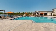 Located at 723 W 19th St in Panama City, Florida, this outdoor amenity space is designed around a generously sized, resort-style swimming pool with crystal-clear blue water and a classic cross-shaped layout. The surrounding pool deck is finished with elegant paver stonework, providing a clean and durable surface throughout. A covered cabana structure along one side of the pool features a flat roof supported by bold columns, ceiling fans for added comfort, and a collection of stylish lounge seating with blue and orange accent furnishings — offering a shaded retreat ideal for relaxation. On the opposite side of the deck, a sun-drenched lounge area is lined with rows of chaise lounges positioned to take full advantage of Florida's abundant sunshine. A secondary pergola structure provides additional shaded seating near the waterfront edge, where a wrought iron fence frames views of the adjacent pond and lush greenery beyond. Vibrant orange market umbrellas are stationed throughout the deck, adding a pop of color and additional sun protection. The multi-story residential buildings visible in the background provide architectural context, while the open sky and natural surroundings contribute to the overall resort-like ambiance of this thoughtfully designed outdoor space.