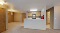 This single-floor residence offers a practical and welcoming open-concept layout designed for comfortable everyday living. The kitchen is positioned behind a peninsula-style breakfast bar that doubles as a natural divider between the cooking and living areas. Light oak cabinetry lines the kitchen walls, providing generous storage space, while stainless steel appliances — including an over-the-range microwave and a freestanding range — add a modern touch. Tile flooring anchors the kitchen area, offering easy maintenance underfoot. The adjoining living area is finished with soft, neutral-toned carpeting that extends throughout, creating a warm and cohesive atmosphere. A textured ceiling runs across the space, complementing the clean, understated wall tones. The open sightlines between the kitchen and living area promote an easy flow and a sense of spaciousness. Interior doors with natural wood finishes provide access to additional rooms, maintaining a consistent aesthetic throughout. The overall design prioritizes functionality and simplicity, making the space well-suited for a variety of living arrangements.