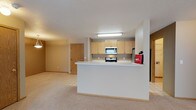 This single-floor residence offers a practical and welcoming open-concept layout designed for comfortable everyday living. The kitchen is positioned behind a peninsula-style breakfast bar that doubles as a natural divider between the cooking and living areas. Light oak cabinetry lines the kitchen walls, providing generous storage space, while stainless steel appliances — including an over-the-range microwave and a freestanding range — add a modern touch. Tile flooring anchors the kitchen area, offering easy maintenance underfoot. The adjoining living area is finished with soft, neutral-toned carpeting that extends throughout, creating a warm and cohesive atmosphere. A textured ceiling runs across the space, complementing the clean, understated wall tones. The open sightlines between the kitchen and living area promote an easy flow and a sense of spaciousness. Interior doors with natural wood finishes provide access to additional rooms, maintaining a consistent aesthetic throughout. The overall design prioritizes functionality and simplicity, making the space well-suited for a variety of living arrangements.