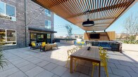 Located at 1725 W Katella Ave in Orange, California, this contemporary outdoor amenity deck offers a thoughtfully designed space for relaxation and social gatherings. The deck is anchored by a striking wood-slatted pergola that provides partial shade while allowing natural light to filter through, creating an inviting interplay of sun and shadow across the large-format stone tile flooring. String lights are strung overhead, adding a warm, festive ambiance during evening hours. A mounted outdoor television and pendant lighting are integrated into the pergola structure, making it well-suited for al fresco entertainment. A generous communal dining table with vibrant yellow accent chairs serves as a central gathering point, while multiple lounge seating areas furnished with stylish outdoor sofas and armchairs invite casual relaxation. A dedicated BBQ grilling station with blue bar seating provides ample space for outdoor cooking and dining. Toward the far end of the deck, a brick fireplace surrounded by cozy lounge chairs creates a warm focal point. Raised planters with desert-inspired landscaping and a backdrop of surrounding residential buildings frame the space, while open skies above enhance the bright, airy Southern California atmosphere throughout.