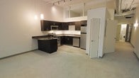 This single-floor space showcases a thoughtfully designed open-concept layout that seamlessly connects the kitchen and living areas. The kitchen is a standout feature, equipped with rich dark cabinetry, granite countertops, and a full suite of stainless steel appliances including a range, microwave, dishwasher, and refrigerator. A peninsula counter extends the workspace and serves as a natural divider between the kitchen and the adjoining carpeted living area, creating a functional yet open flow throughout the space. Soaring ceilings with exposed structural beams lend an airy, loft-like quality to the interior. Overhead track lighting and pendant lights illuminate the kitchen with a warm, modern glow, while large white structural columns add architectural character. The flooring transitions from tile in the kitchen to plush carpeting in the living area, offering both practicality and comfort. A white interior door leads to an additional room, suggesting private quarters beyond the open living space. The overall design balances contemporary finishes with industrial-inspired elements, resulting in a clean, modern aesthetic that is both functional and visually appealing.