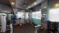 This fitness center offers a functional and well-appointed workout environment designed to support a range of exercise routines. The space is outfitted with a cable crossover machine as its centerpiece, accompanied by additional strength-training equipment including a seated rowing machine and an adjustable weight bench, providing users with versatile options for both upper and lower body workouts. Additional equipment is visible in the mirrored reflection, suggesting a broader selection throughout the space. The flooring features durable dark rubber matting, ideal for high-use gym environments. Mirrored walls line the perimeter, visually expanding the space and allowing users to monitor their form during exercise. Multiple windows fitted with horizontal blinds allow natural light to filter in, creating a bright and open atmosphere. Ceiling fans are mounted overhead, contributing to air circulation and comfort during workouts. Recessed lighting provides consistent illumination throughout. The teal accent wall adds a pop of color, lending the space a modern, energetic aesthetic that complements its practical design.