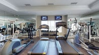 This fitness center offers a thoughtfully arranged workout space designed to accommodate a variety of exercise routines. The room features hardwood-style flooring and a clean, neutral color palette with white walls and crown molding, lending the space a polished, welcoming atmosphere. Natural light filters in through multiple windows fitted with horizontal blinds, providing a bright environment while maintaining privacy.

The cardio area is prominently positioned at the center of the room, housing multiple treadmills and an elliptical trainer. Two wall-mounted flat-screen televisions are positioned above the windows, allowing users to stay entertained during cardio sessions. A well-stocked dumbbell rack lines the back wall, offering a range of free weights to support strength training.

Floor-to-ceiling mirrors are installed along the side walls, visually expanding the space and allowing users to monitor their form during workouts. Additional strength and resistance equipment is visible along the perimeter, making the facility suitable for full-body training. Ceiling-recessed lighting supplements the natural light, ensuring consistent illumination throughout.