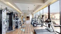 This fitness center is organized into two distinct zones designed to support a comprehensive workout experience. The main exercise area features an extensive collection of cardio equipment, including treadmills, elliptical trainers, stationary bikes, and cable machines, all arranged along a wall of floor-to-ceiling windows that provide direct views of an outdoor pool and surrounding landscape. Natural light floods the space throughout the day, complemented by recessed and cove lighting in the coffered ceiling. A ceiling fan overhead aids in air circulation, and large mirrors along the walls visually expand the room. Dumbbell racks and multiple strength-training machines are positioned throughout, offering a full range of resistance options. A wall-mounted television is centrally placed for entertainment during workouts. The decorative tile flooring features a circular pattern that adds visual interest underfoot. Adjacent to the main floor is a dedicated studio room with soaring ceilings, hardwood flooring, and curved mirror walls that reflect abundant natural light from clerestory windows above. Wooden shelving units store yoga mats, stability balls, and other fitness accessories, making this studio well-suited for group classes or individual flexibility and balance training.